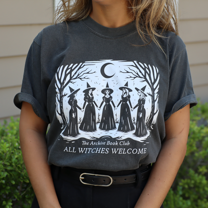 A person wearing a dark charcoal gray graphic t-shirt with a white illustration of five witches under a crescent moon and the text 'The Archive Book Club ALL WITCHES WELCOME'. The background shows green bushes and a beige house exterior.