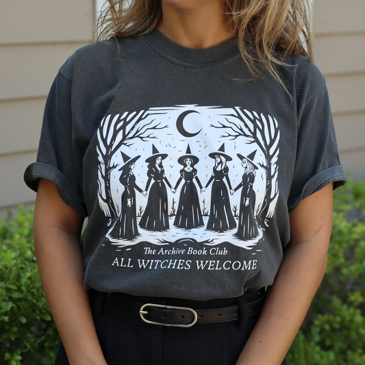 A person wearing a dark charcoal gray graphic t-shirt with a white illustration of five witches under a crescent moon and the text 'The Archive Book Club ALL WITCHES WELCOME'. The background shows green bushes and a beige house exterior.