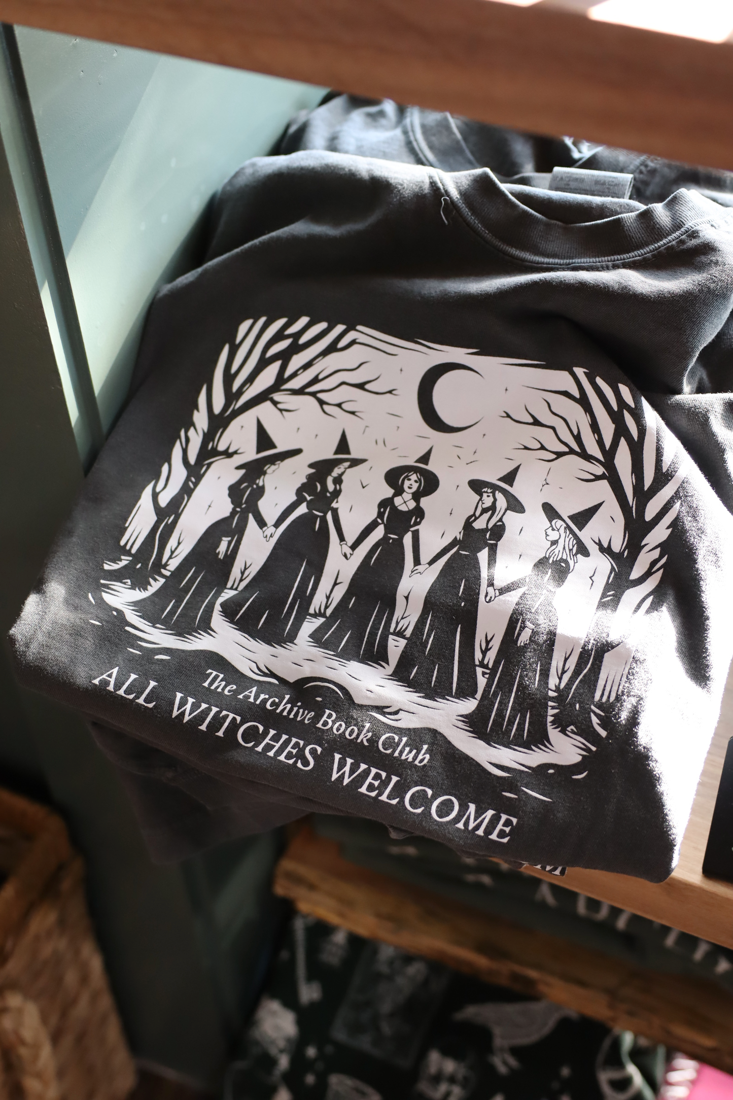 A black graphic t-shirt displayed on a wooden shelf, featuring a white illustration of four witches in hats under a crescent moon with the text 'The Archive Book Club ALL WITCHES WELCOME'.