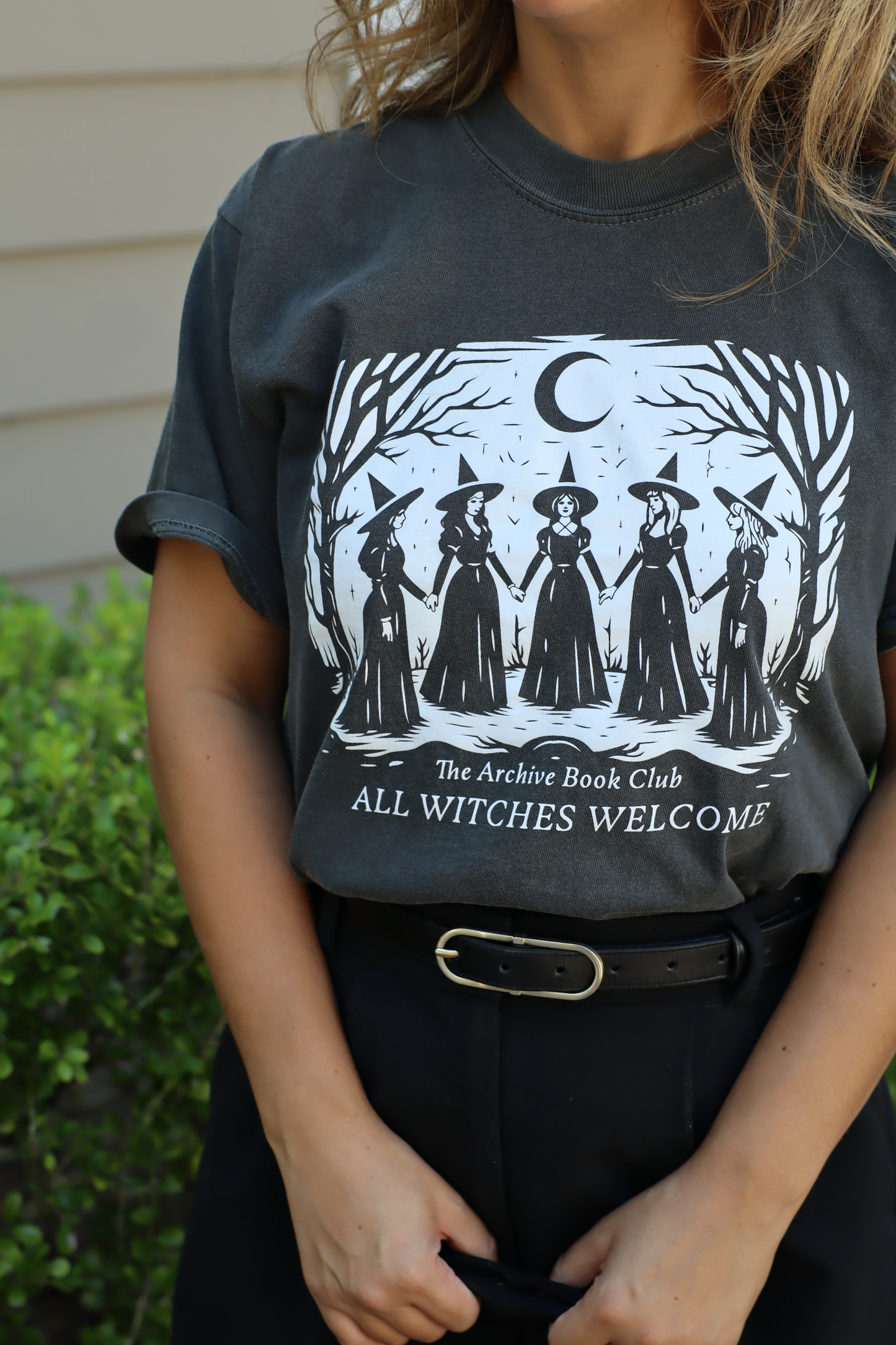 A person wearing a dark charcoal gray graphic t-shirt with a black and white illustration of five witches under a crescent moon and the text 'The Archive Book Club ALL WITCHES WELCOME'.