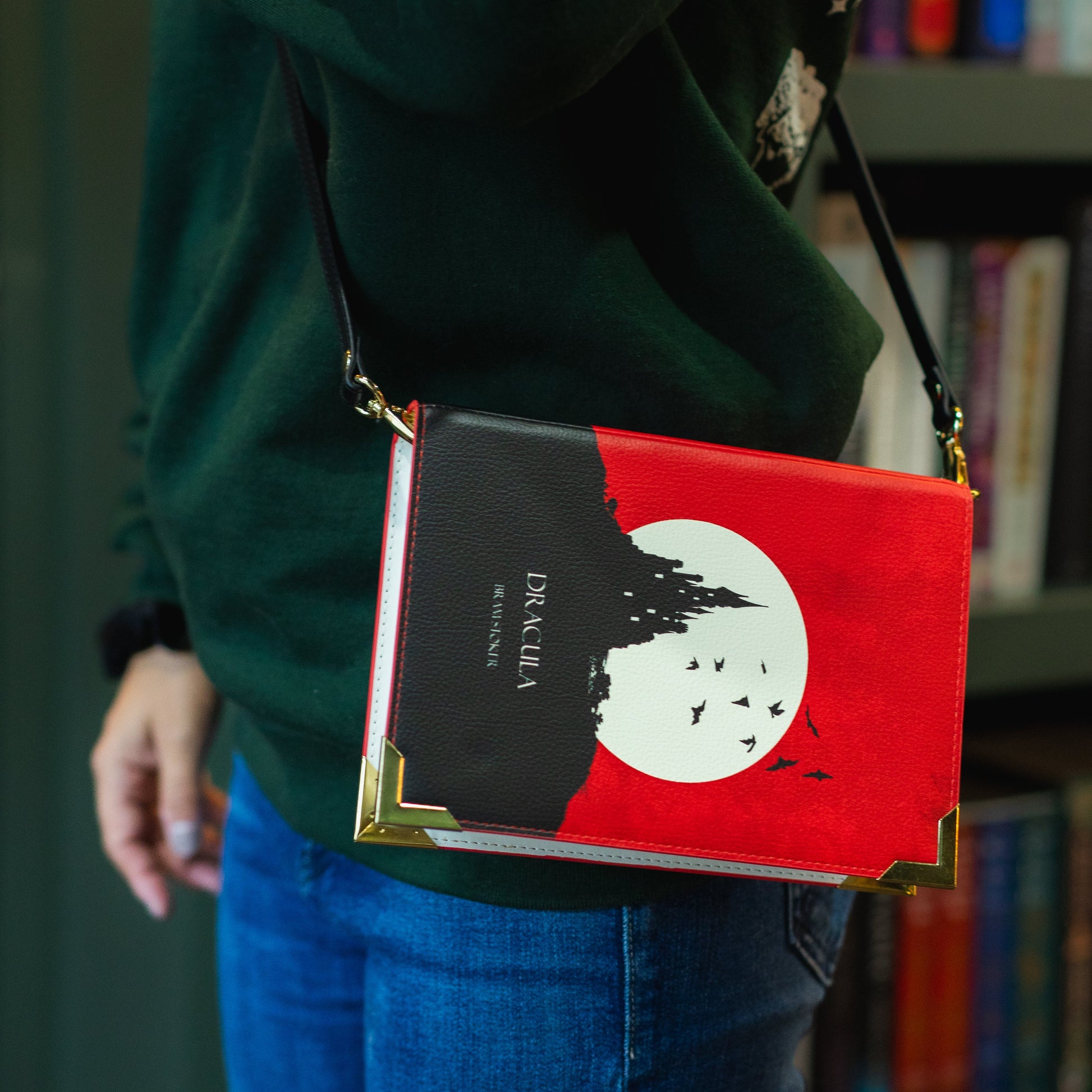 Person wearing a crossbody bag with a book design, standing in front of a bookshelf.