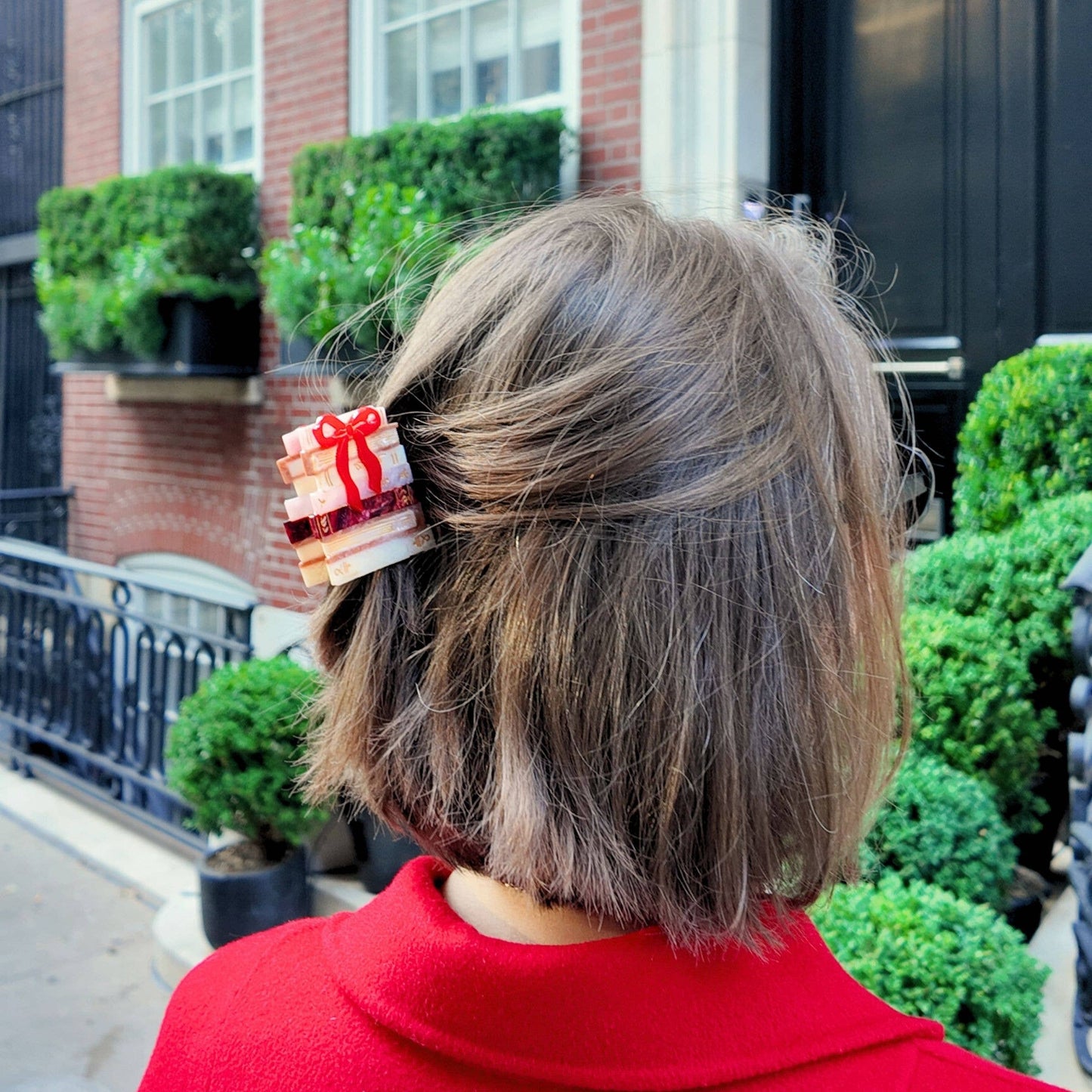 Person with a decorative hair clip in a red coat standing in front of a brick building with greenery.