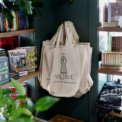 Beige tote bags with 'The Archive' logo hanging on a dark green wall next to bookshelves.