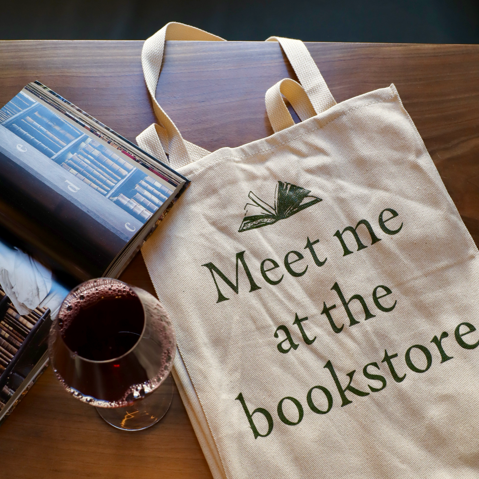 Tote bag with 'Meet me at the bookstore' text on a wooden table with books and a cup.