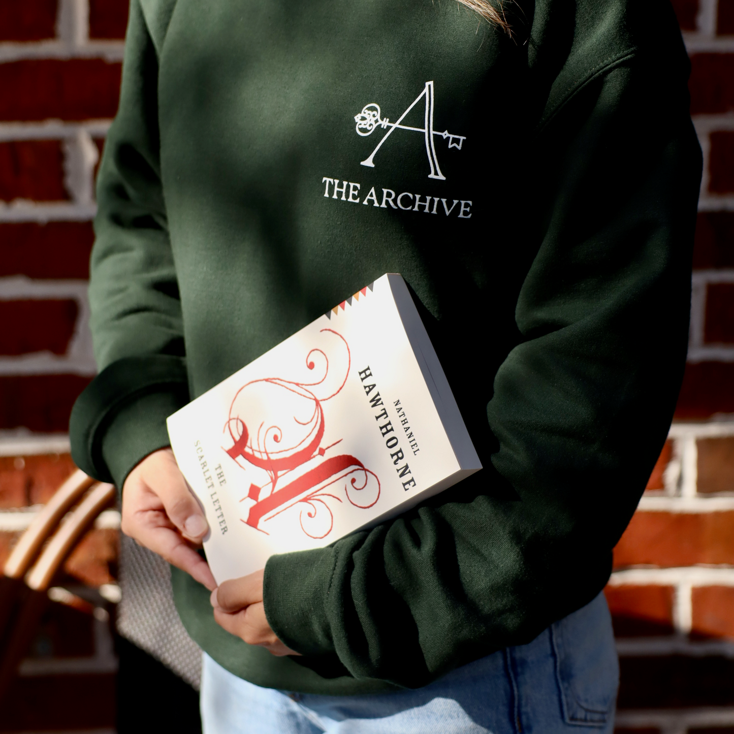 Person wearing a green sweatshirt with 'The Archive' logo, holding a book against a brick wall.