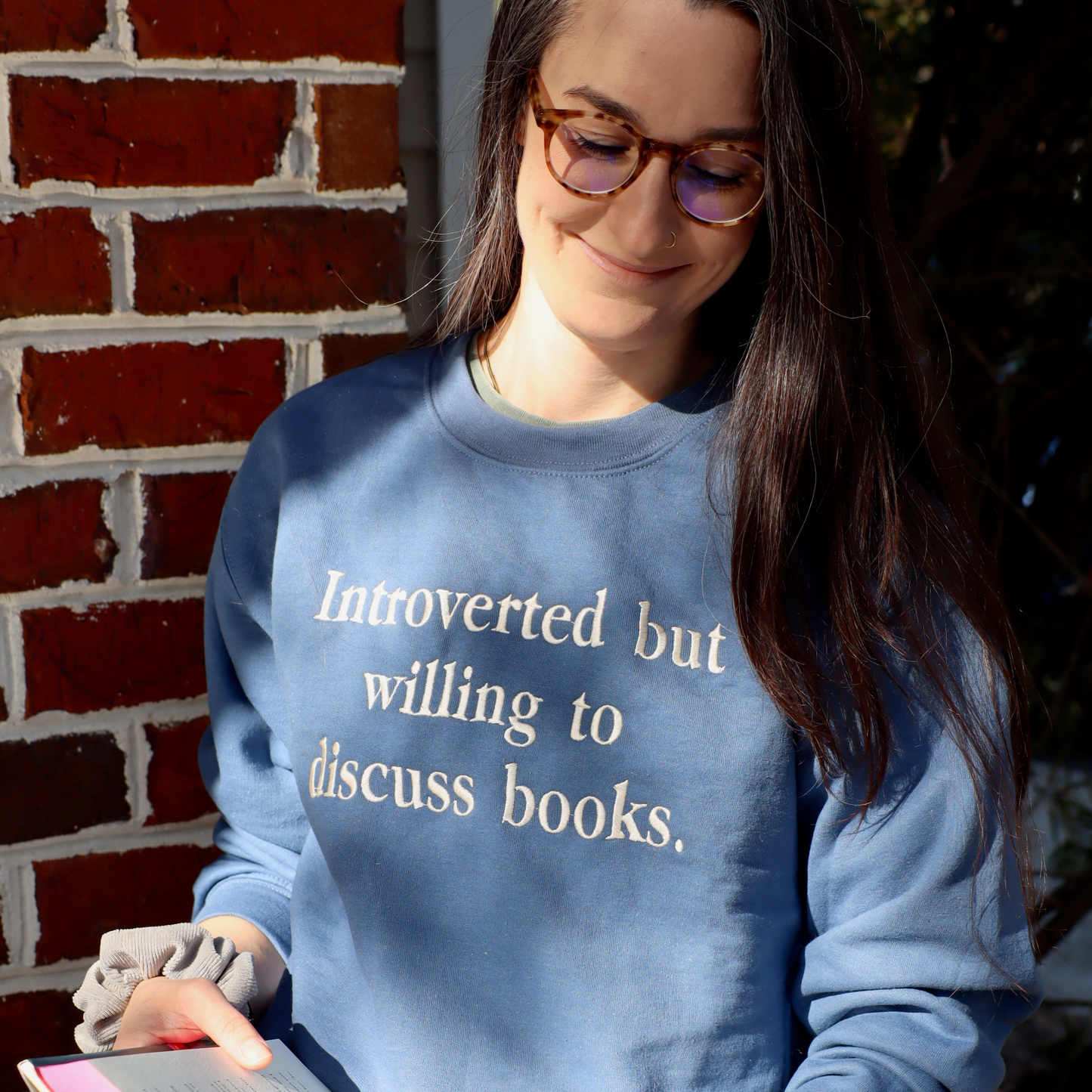 Woman wearing a blue sweatshirt with text, holding a book against a brick wall.