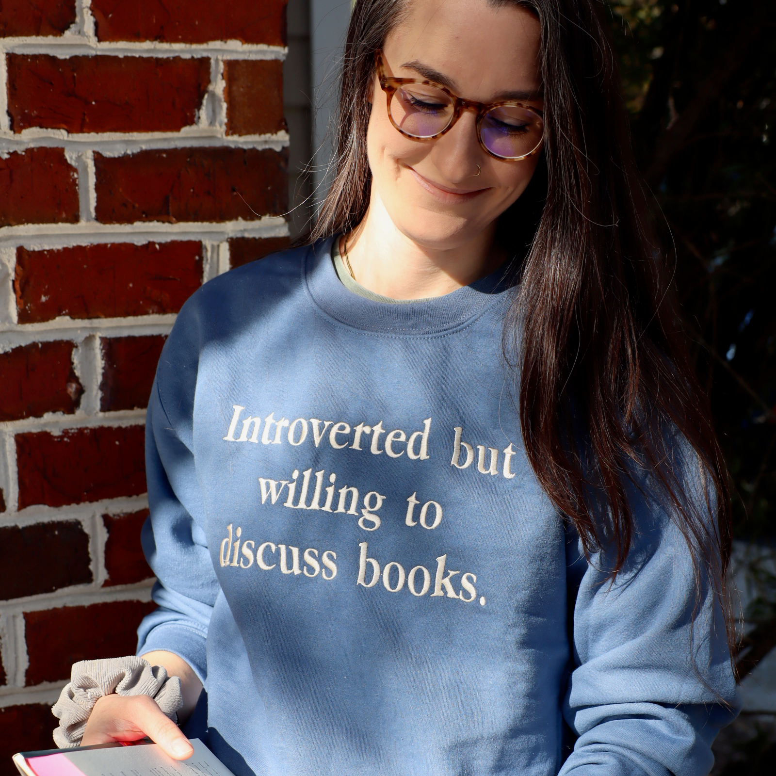 Woman wearing a blue sweatshirt with text, holding a book against a brick wall.