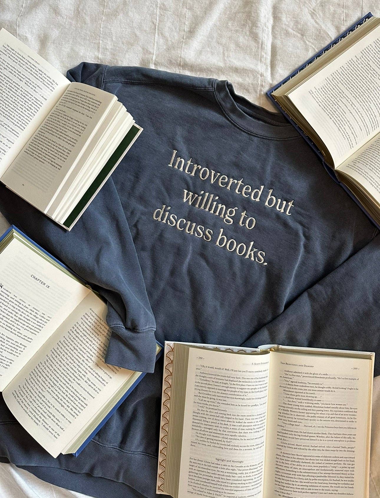 A gray crewneck sweatshirt with the text 'Introverted but willing to discuss books.' printed on the front, laid flat with open books around it.