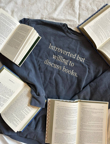 A gray crewneck sweatshirt with the text 'Introverted but willing to discuss books.' printed on the front, laid flat with open books around it.