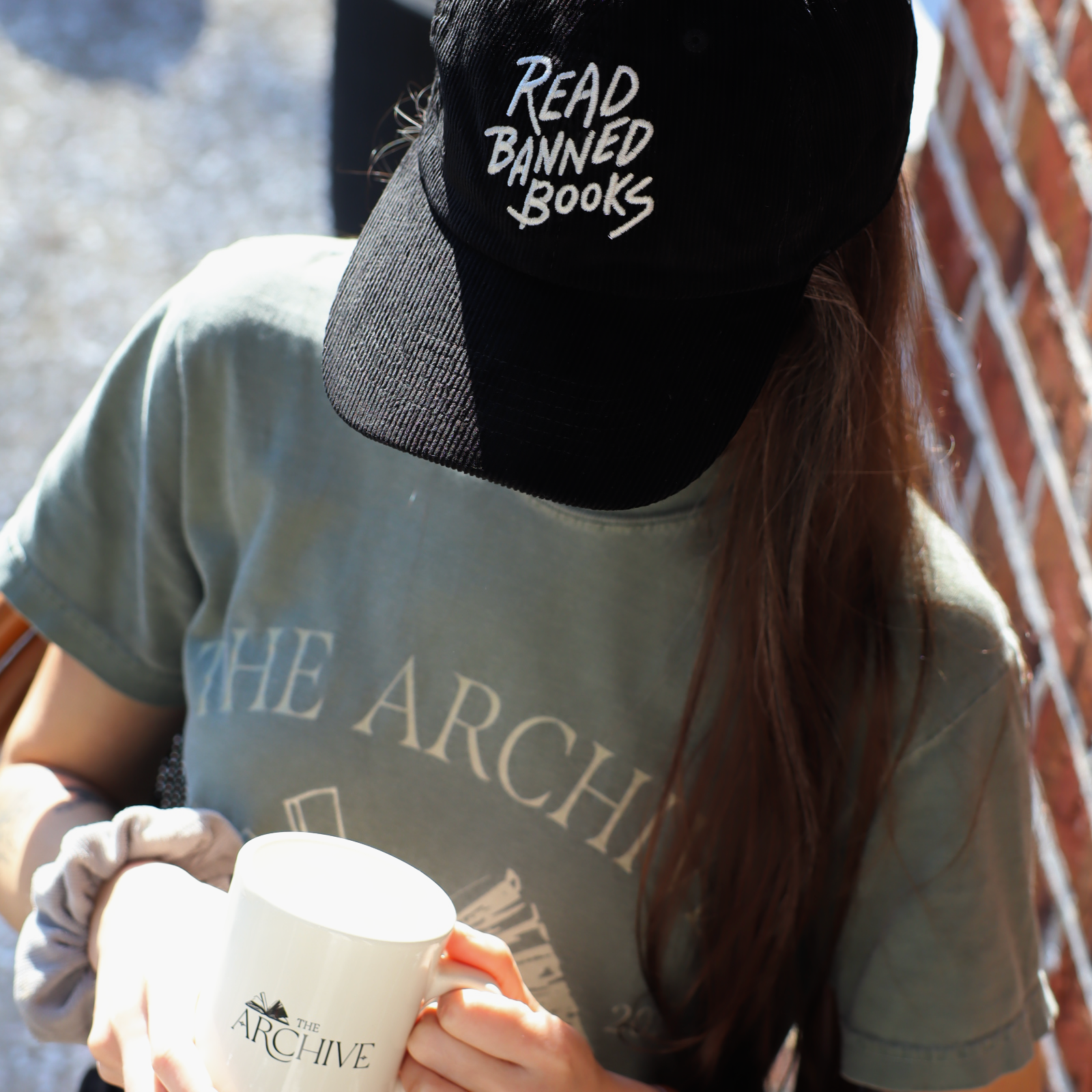 Person wearing a cap with 'Read Banned Books' text, holding a mug with 'The Archive' branding.