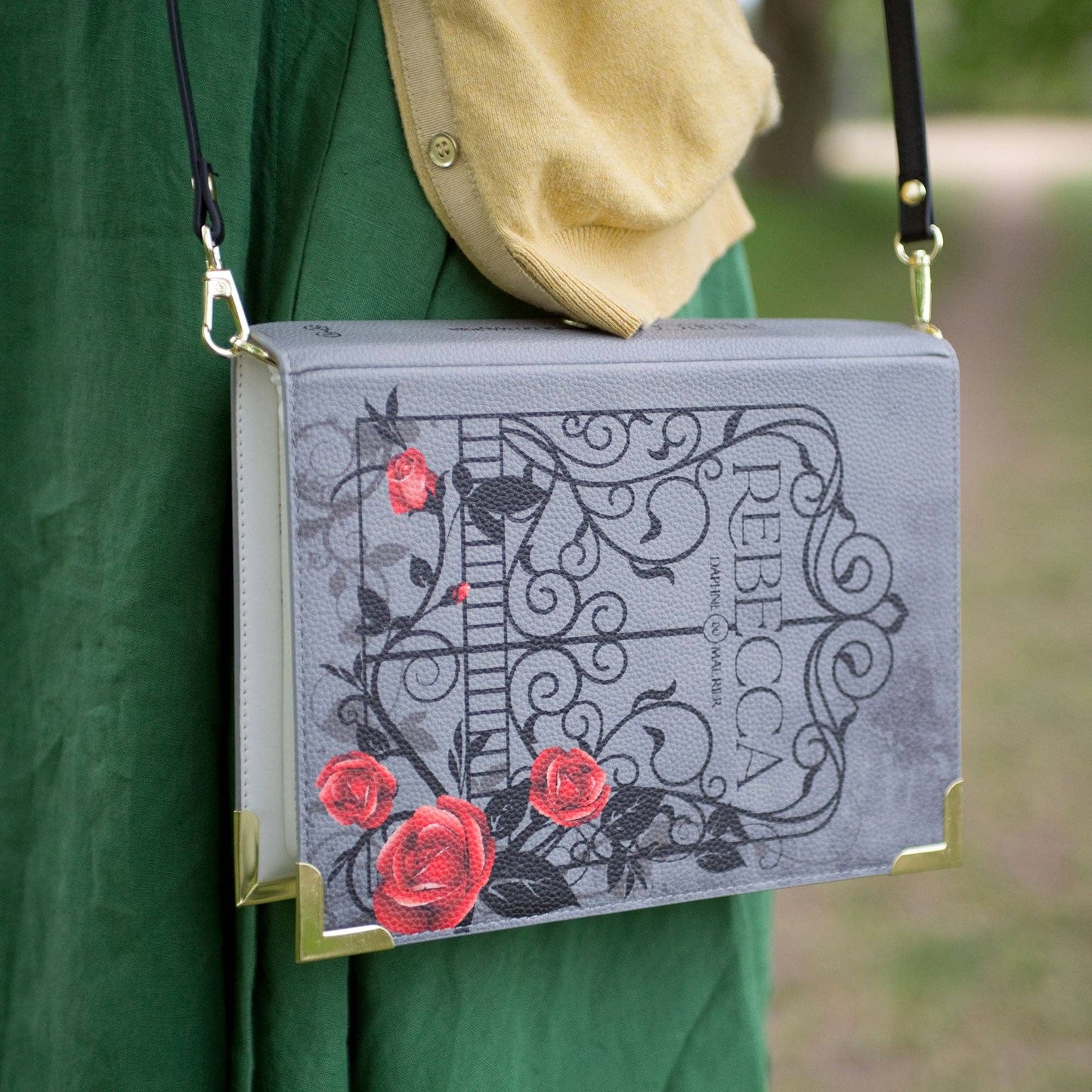 Book-shaped handbag with floral design and 'Rebecca' text, held by a person outdoors.