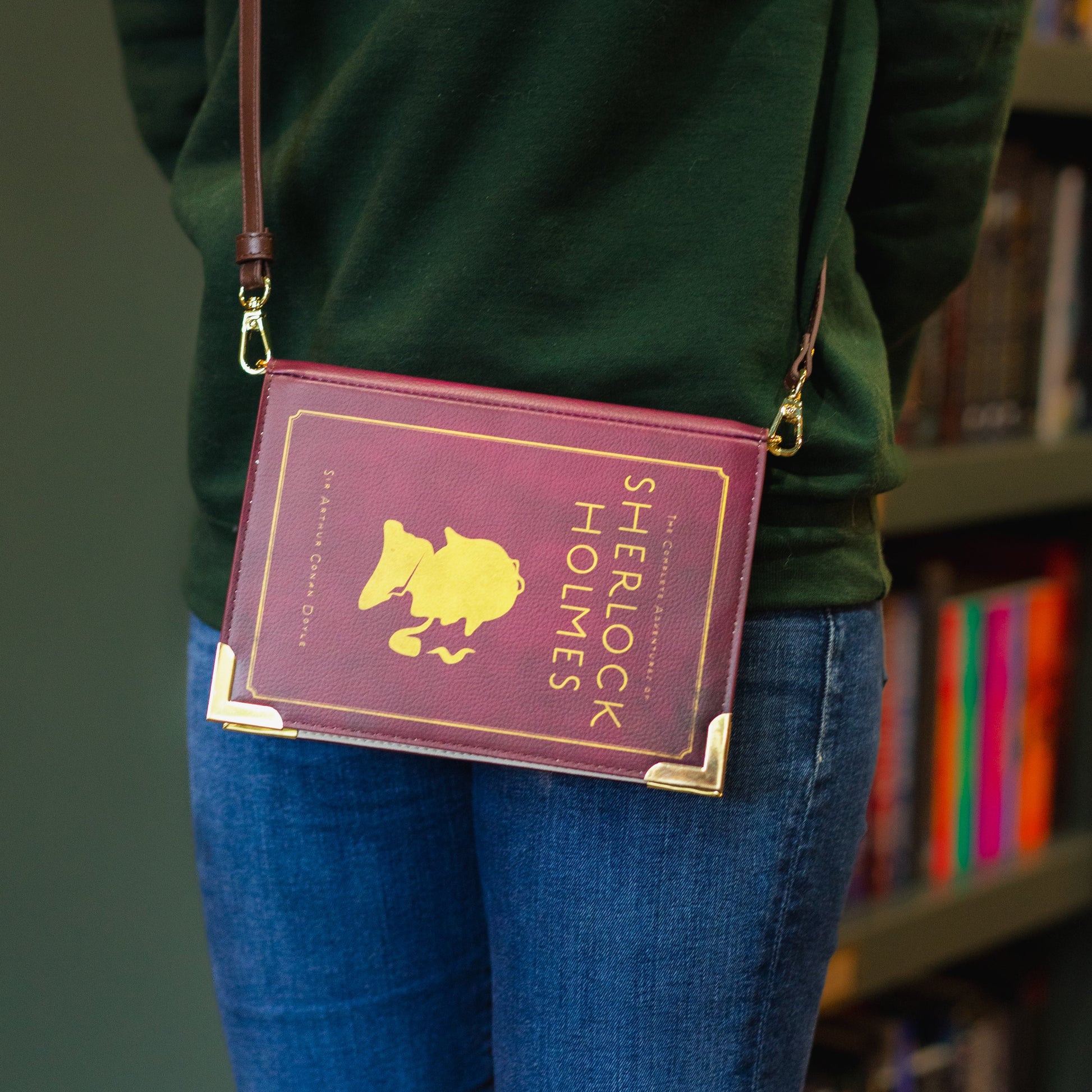Person wearing a green sweatshirt with 'The Archive' logo, holding a purple book-shaped bag with Sherlock Holmes design.