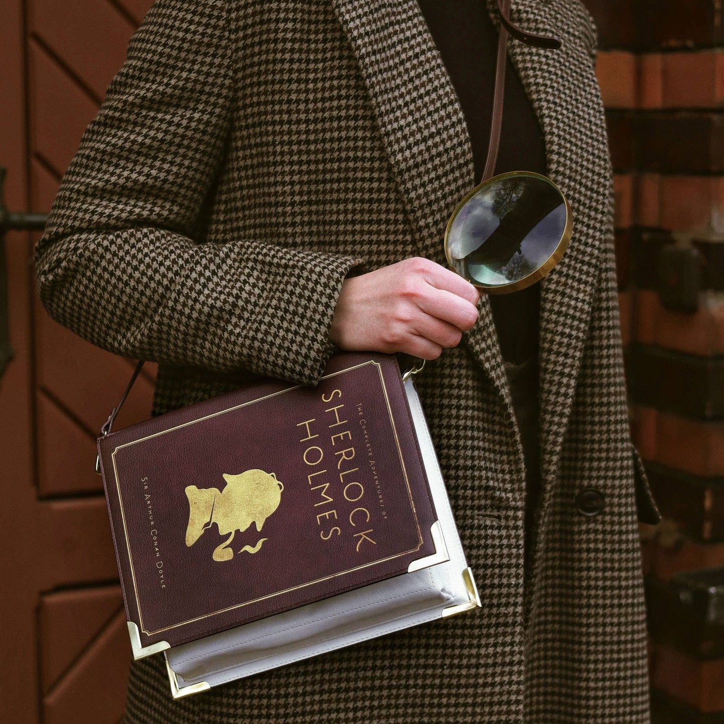 Person in a coat holding a book titled 'Sherlock Holmes' and a magnifying glass against a brick wall.