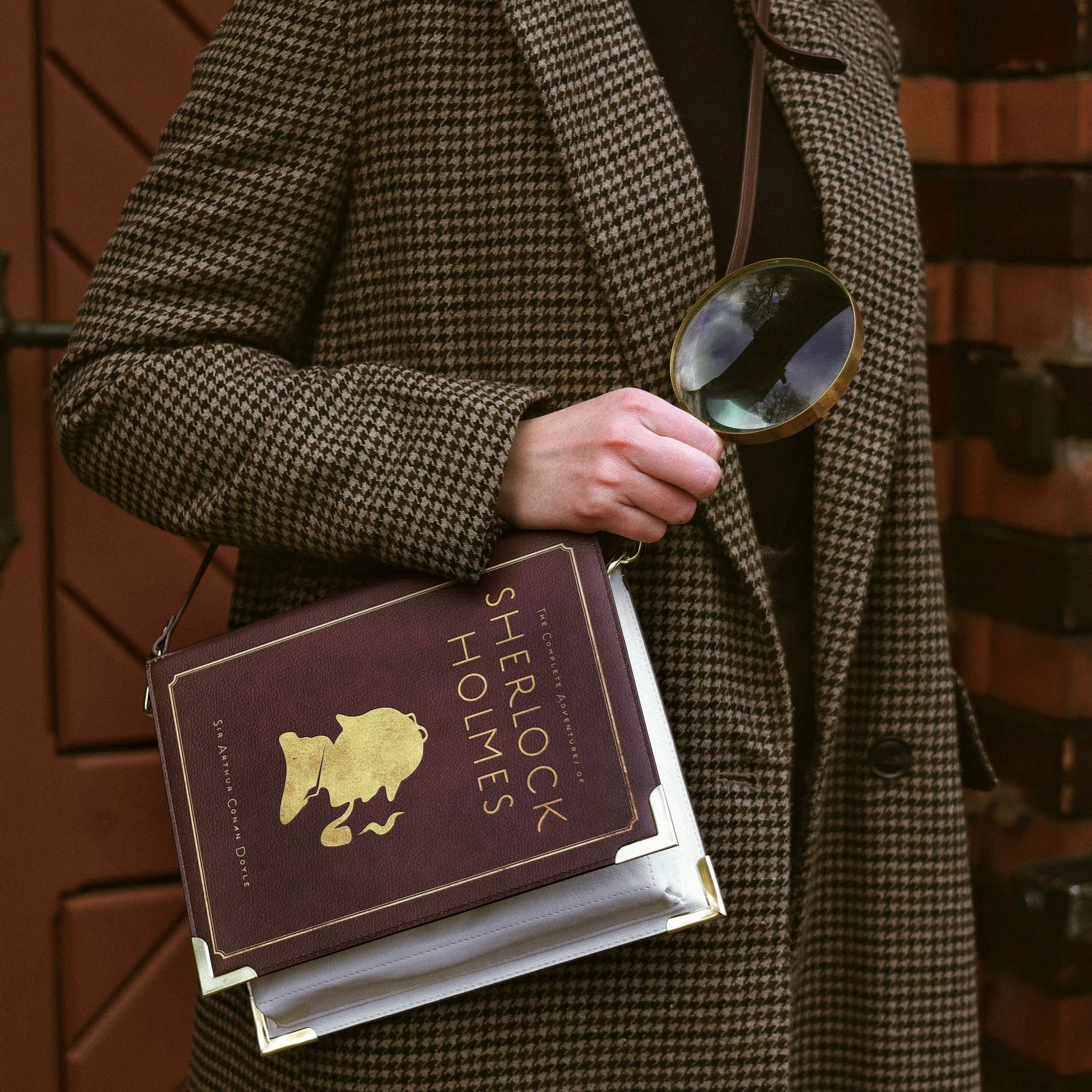 Person in a coat holding a book titled 'Sherlock Holmes' and a magnifying glass against a brick wall.