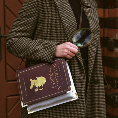 Person in a coat holding a book titled 'Sherlock Holmes' and a magnifying glass against a brick wall.