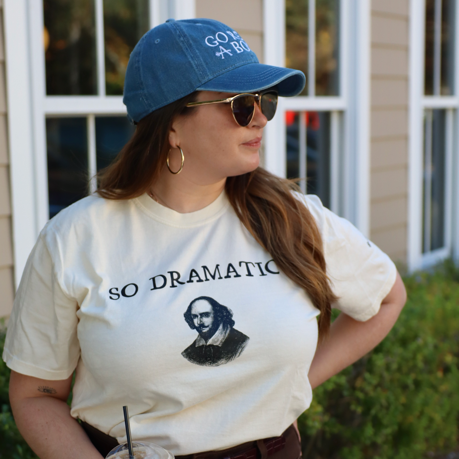 Person wearing a white t-shirt with a graphic and text, holding a drink, and wearing a blue cap.