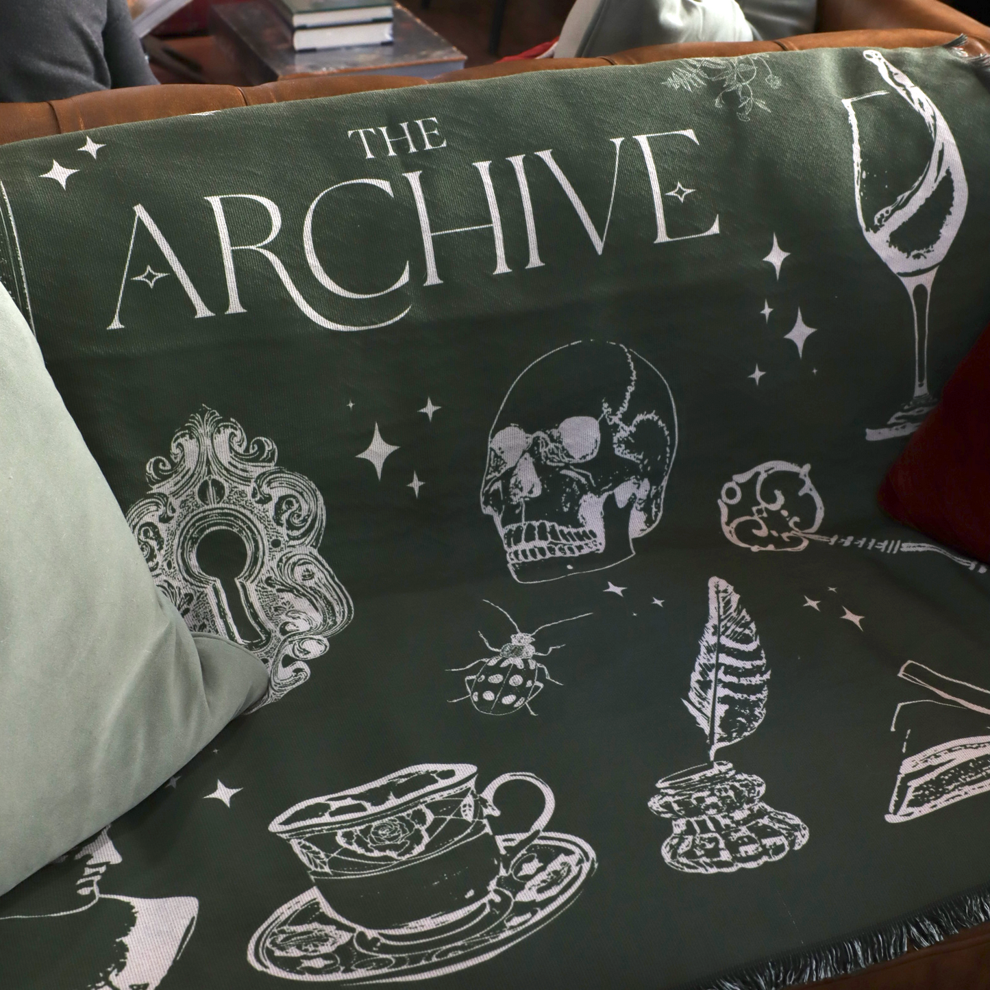 Sofa with a green The Archive Blanket featuring white illustrations and 'The Archive' text.
