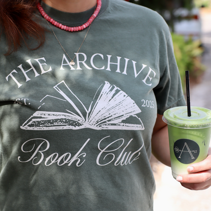 Person wearing a t-shirt with 'The Archive Book Club' design, holding a green drink.