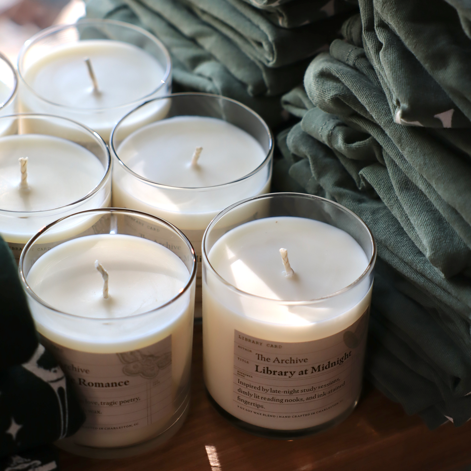 Candles in glass jars with labels on a wooden surface, surrounded by green folded shirts.