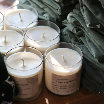 Candles in glass jars with labels on a wooden surface, surrounded by green folded shirts.
