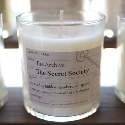 Candle in a glass jar with a label titled 'The Secret Society' on a wooden surface.