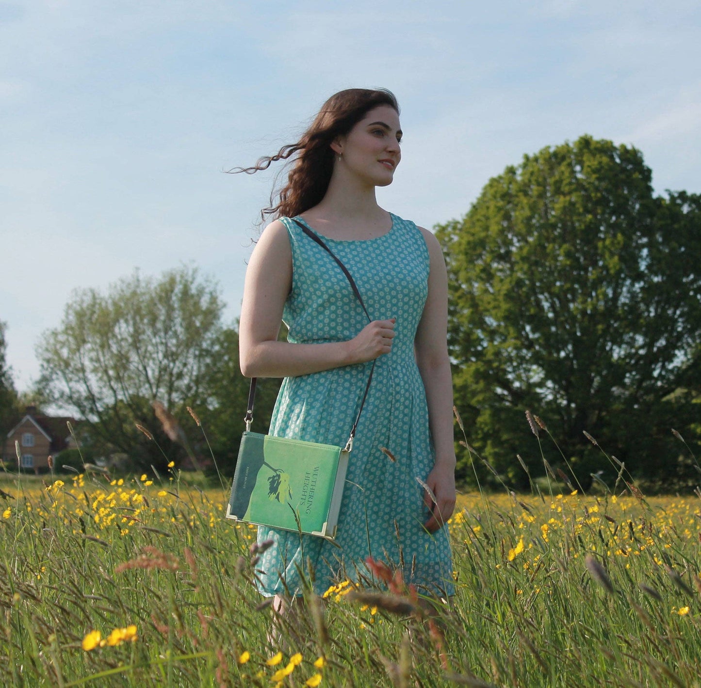 Woman in a green dress holding a green Wuthering Heights Green Book Crossbody Handbag Clutch  in a field with yellow flowers and trees in the background.